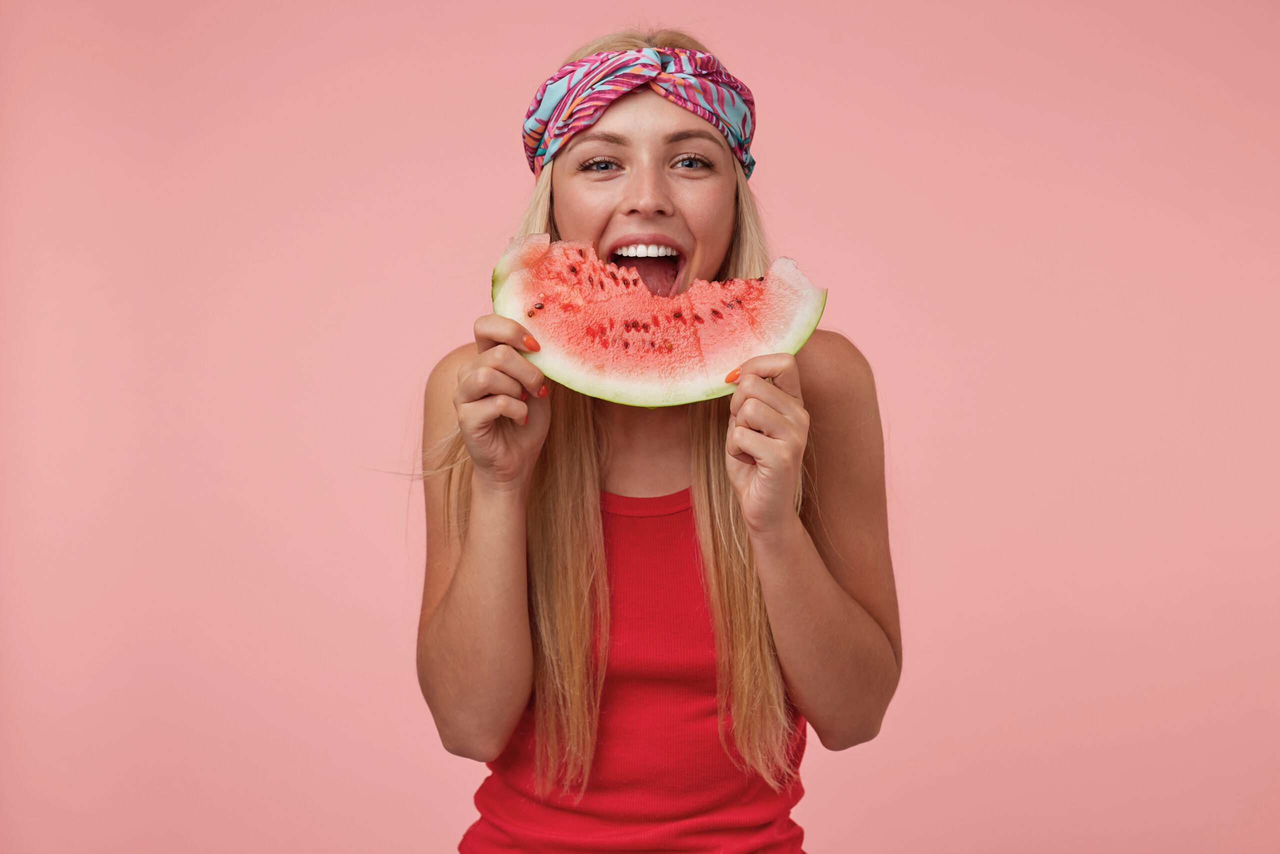 Indoor shot of joyful pretty female with headband and long blonde hair posing over pink background, eating watermelon, being in nice mood