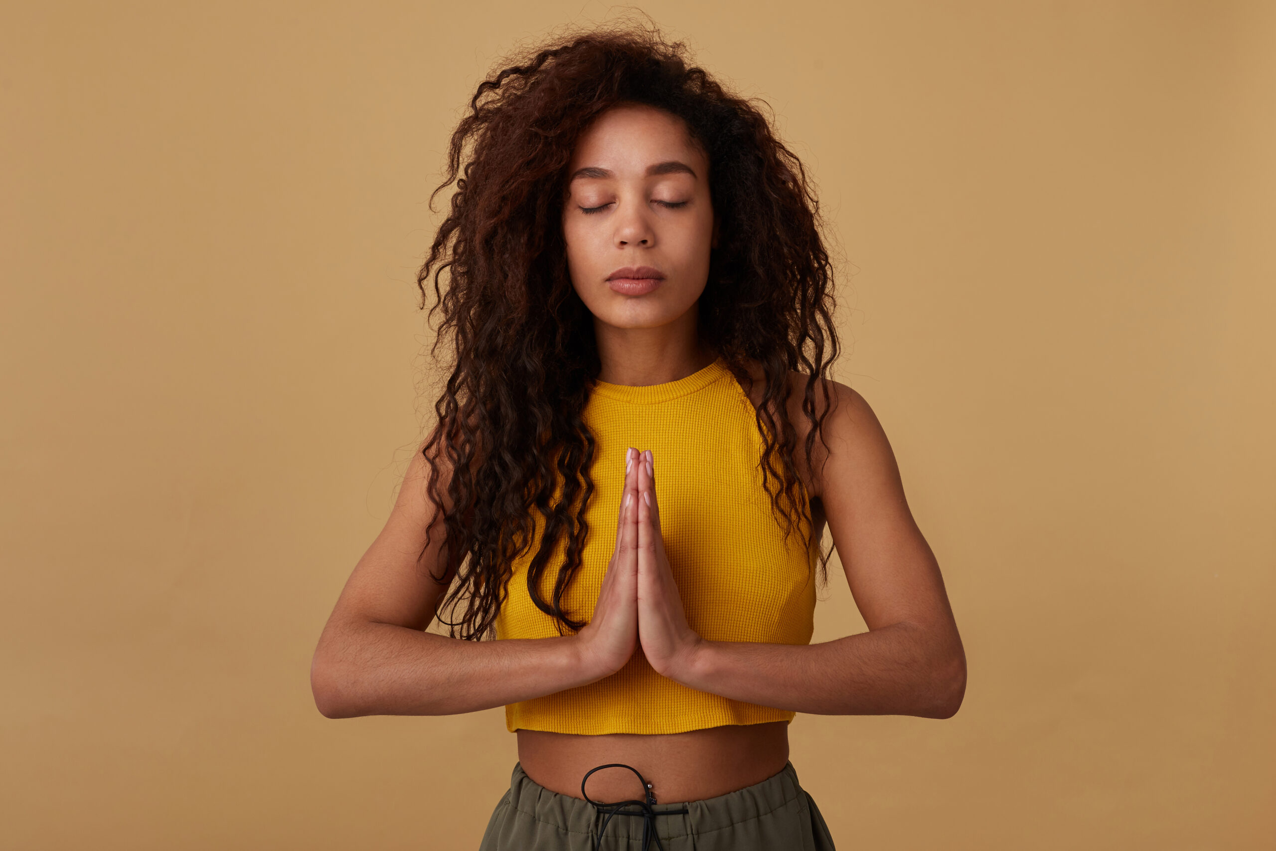 Pleasant looking young calm curly brunette dark skinned woman keeping her eyes closed while meditating and raising folded hands, isolated over beige background