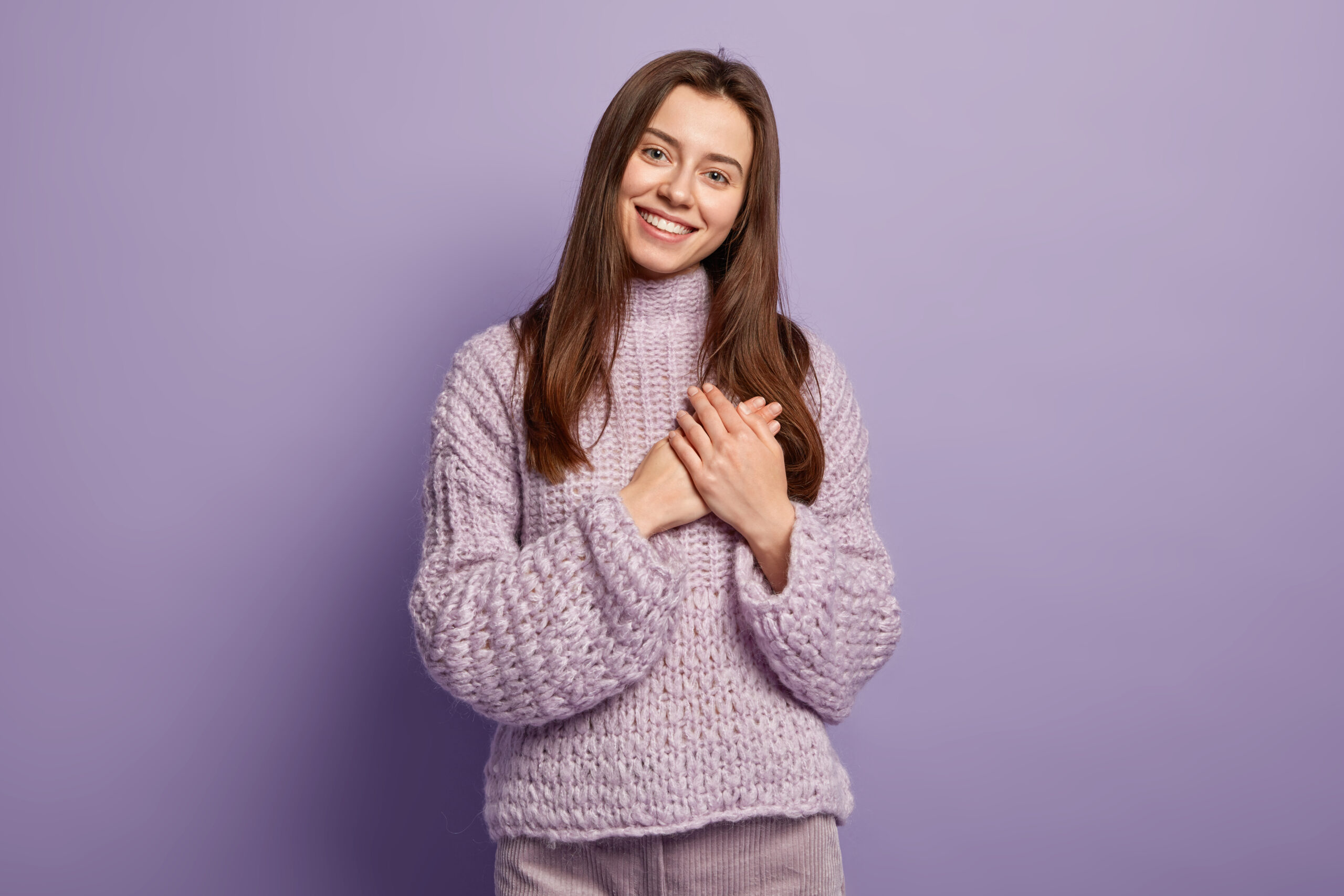 Portrait of happy young European woman keeps hands on breast, shows heart gesture, expresses gratitude, being thankful, models against purple background. Body language. Monochrome. People and devotion