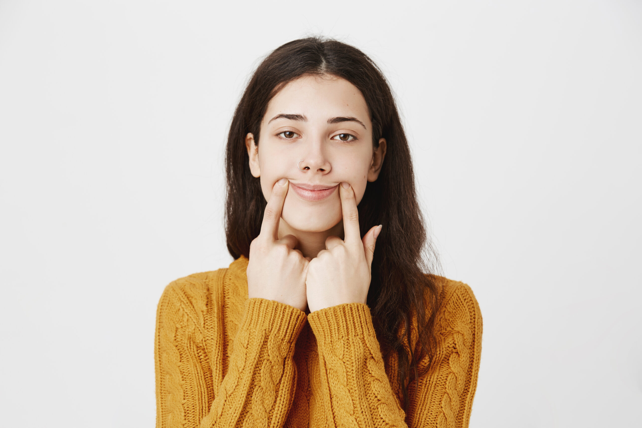 Close-up portrait of tired and gloomy caucasian pierced girl stretching mouth with fingers, making fake smile while feeling sad, standing over gray background. Woman is not in good mood.