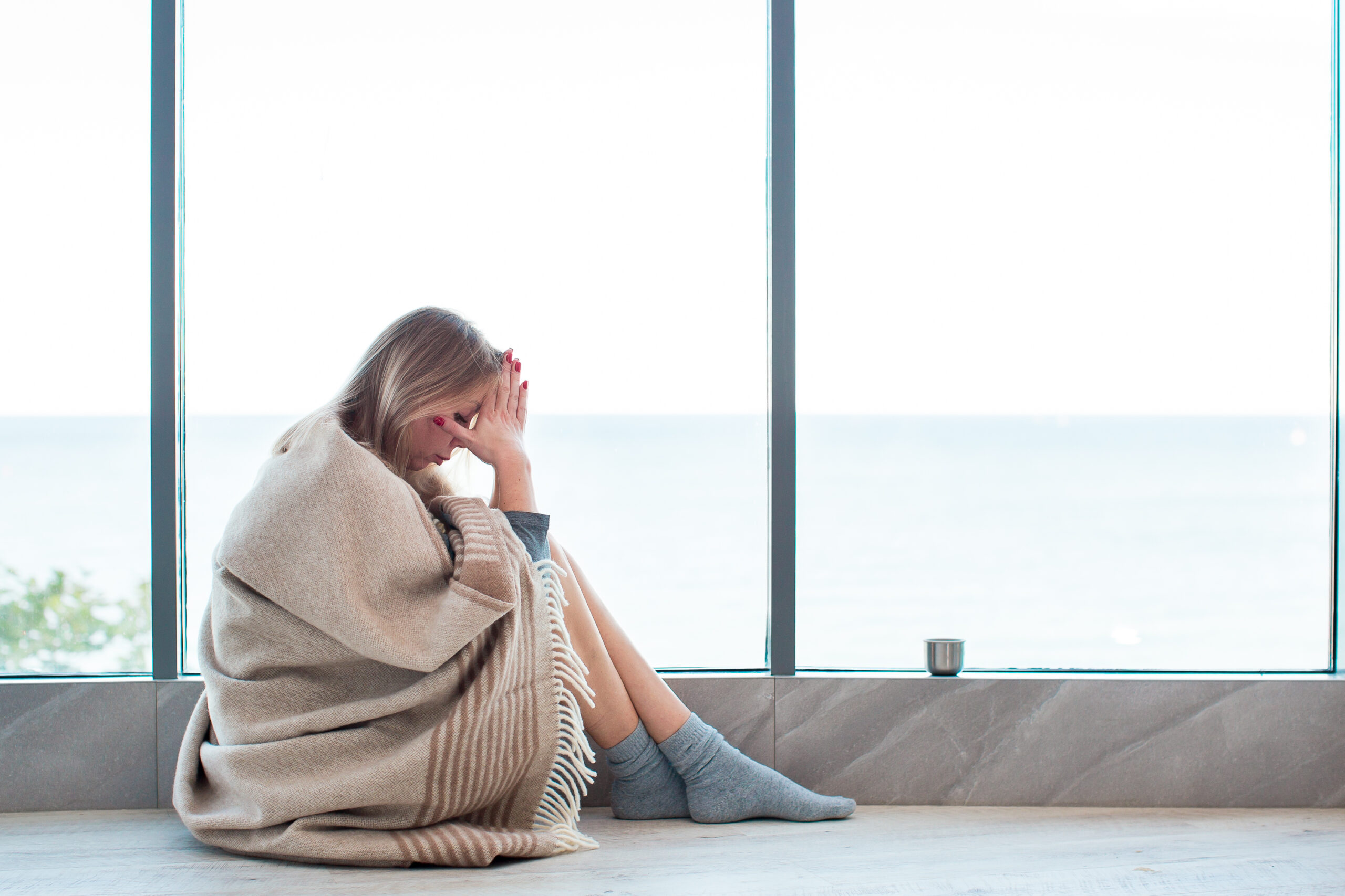 Woman in warm socks sitting on a floor near large window wrapped in a blanket, holding her head, having a strong headache.